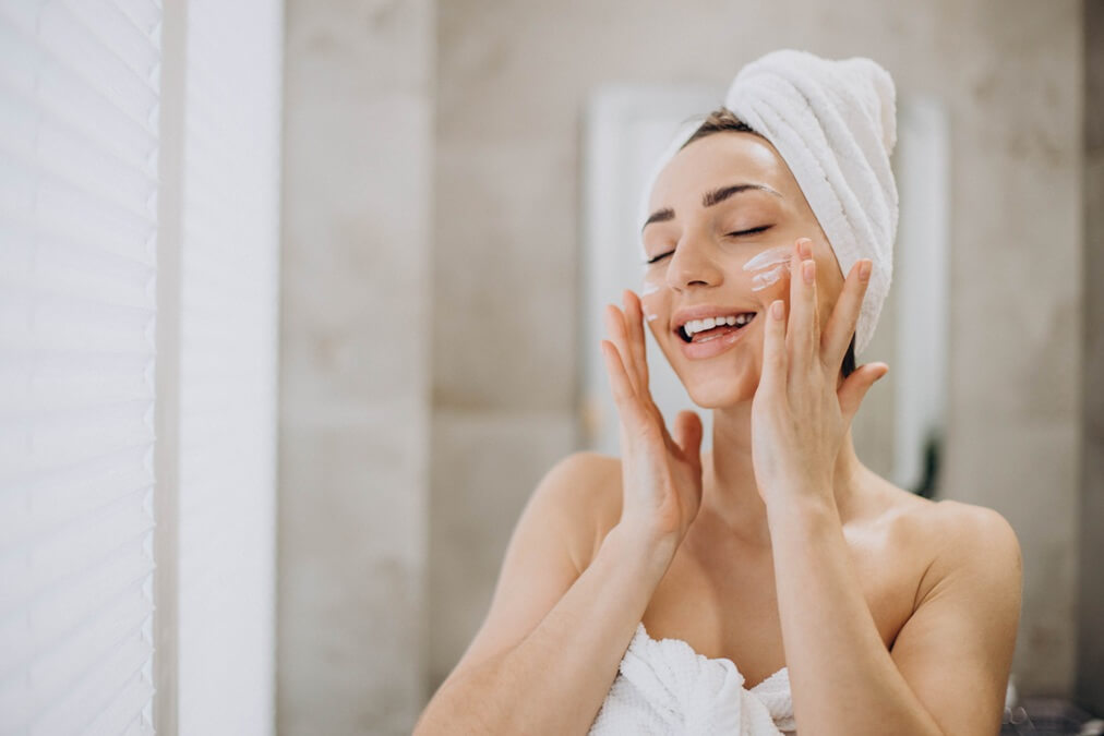 young woman applying face cream her face with towel head 1
